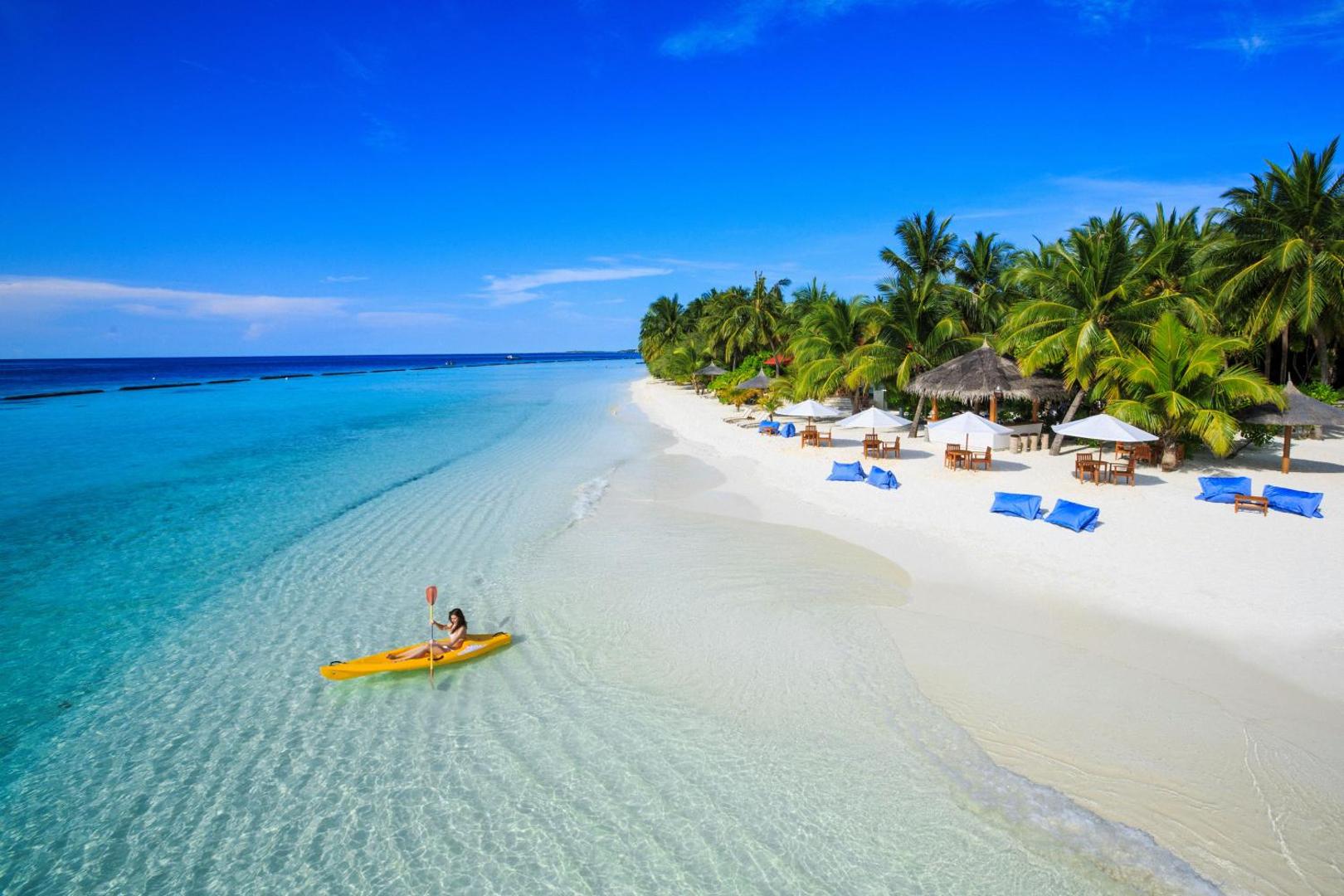 Niva Kurumba Maldives pludmales josla ar atpūtas krēsliem, saulessargiem un tūristu dzeltenā kajakā dzidrā, seklā piekrastes ūdenī