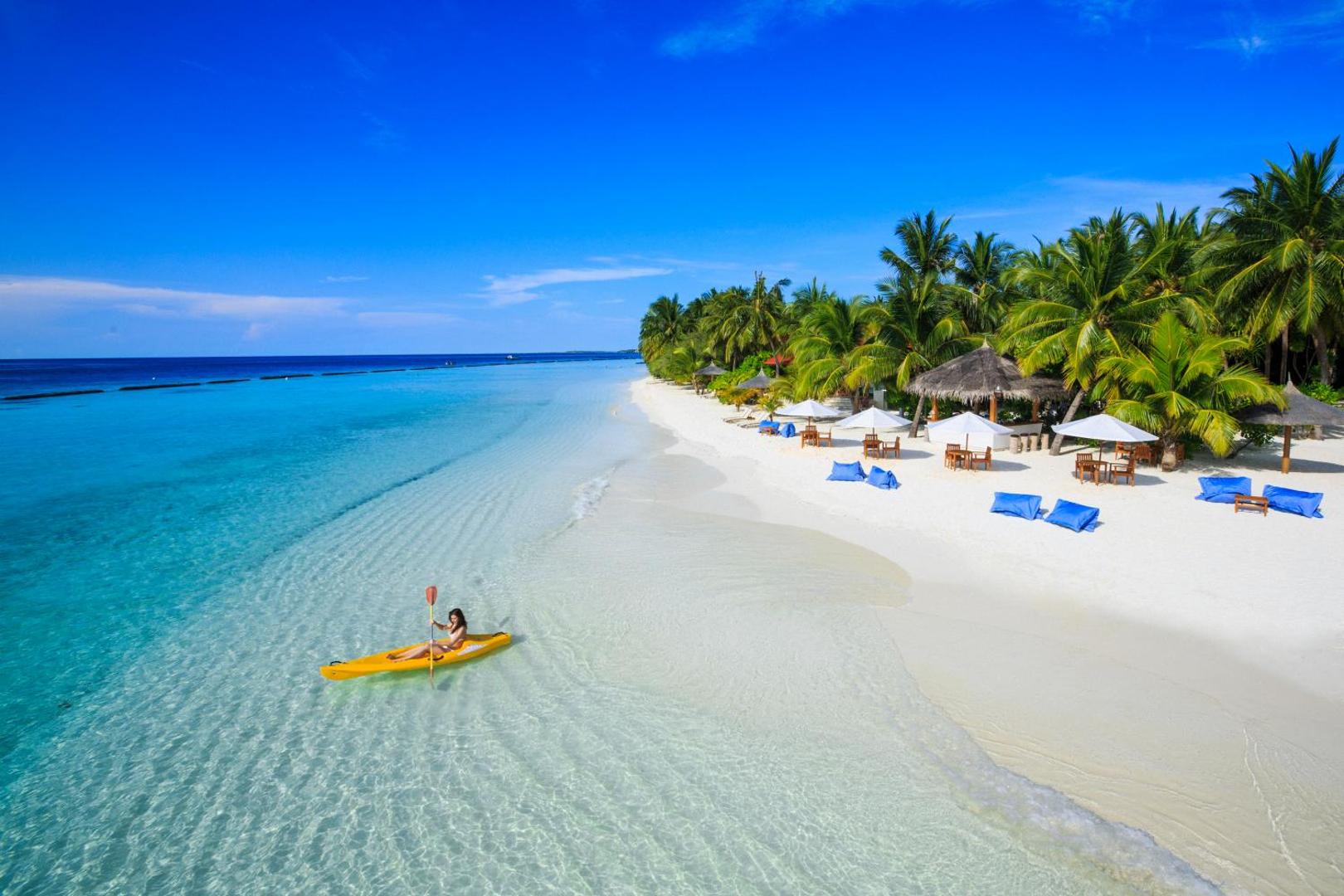 Niva Kurumba Maldives kūrorta pludmale ar baltām smiltīm, tirkīza ūdeni un personu dzeltenā kajakā piekrastes joslā