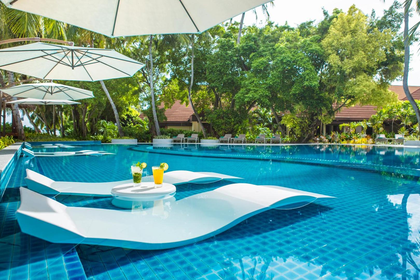 Niva Kurumba Maldives āra baseins ar moderniem sauļošanās krēsliem ūdenī, kokteiļiem un baltiem saulessargiem tropu dārzā.