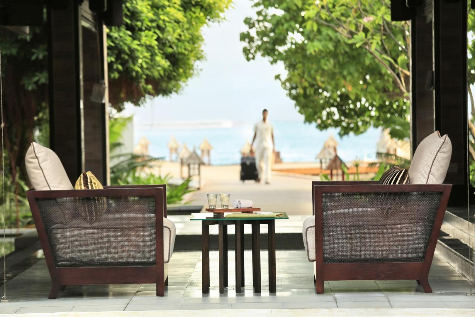 Kurumba Maldives viesnīcas atpūtas zona ar skatu uz okeānu, kurā redzami divi pīti krēsli un darbinieks ar bagāžu fonā