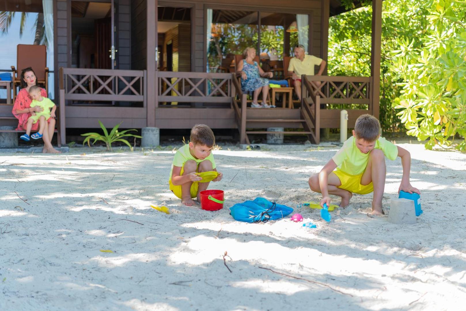Ģimene atpūšas pie koka bungalo Royal Island Resort & Spa — bērni spēlējas smiltīs, kamēr pieaugušie sēž uz terases
