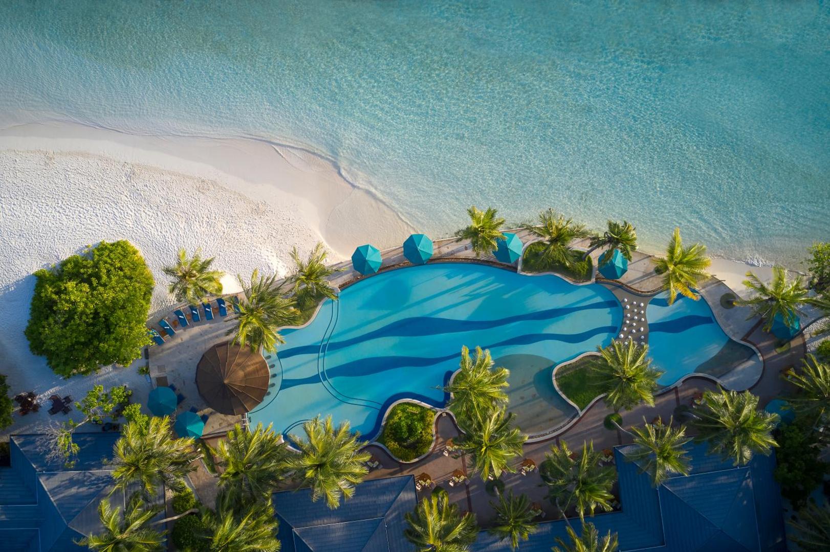 Royal Island Resort & Spa skats no augšas uz galveno baseinu pie pludmales Maldivu salās — palmas, zili saulessargi un tirkīza ūdens