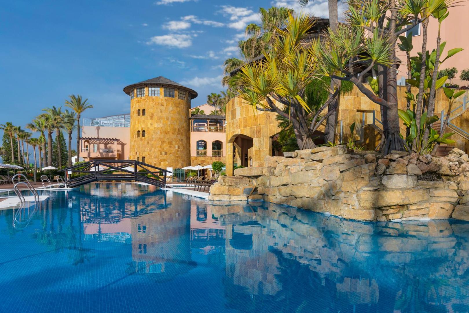 Elba Estepona Gran Hotel And Thalasso Spa arhitektūra — akmens torņi, baseins ar koka tiltiņu un eksotiski augi saulainā dienā