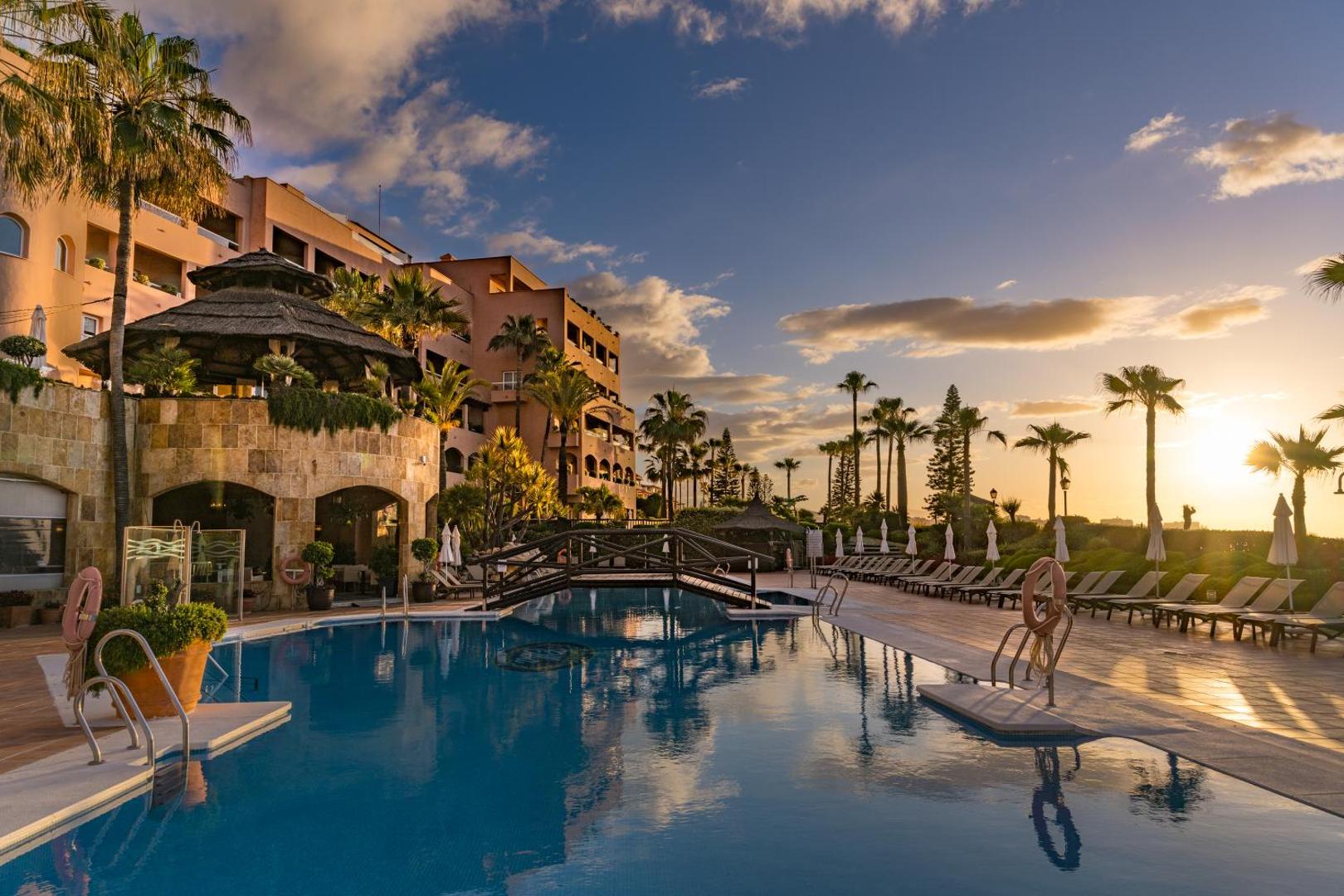 Elba Estepona Gran Hotel And Thalasso Spa āra baseins saulrietā — skats uz viesnīcas fasādi, palmu dārzu un koka tiltiņu
