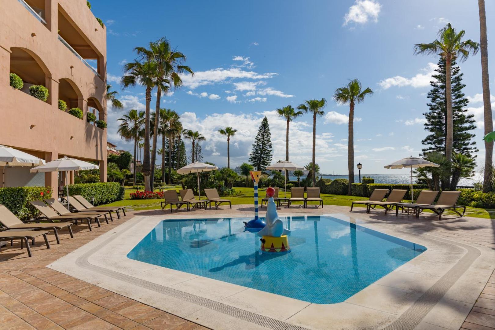 Elba Estepona Gran Hotel And Thalasso Spa bērnu baseins pie viesnīcas ēkas ar zālāju, palmām un sauļošanās krēsliem.