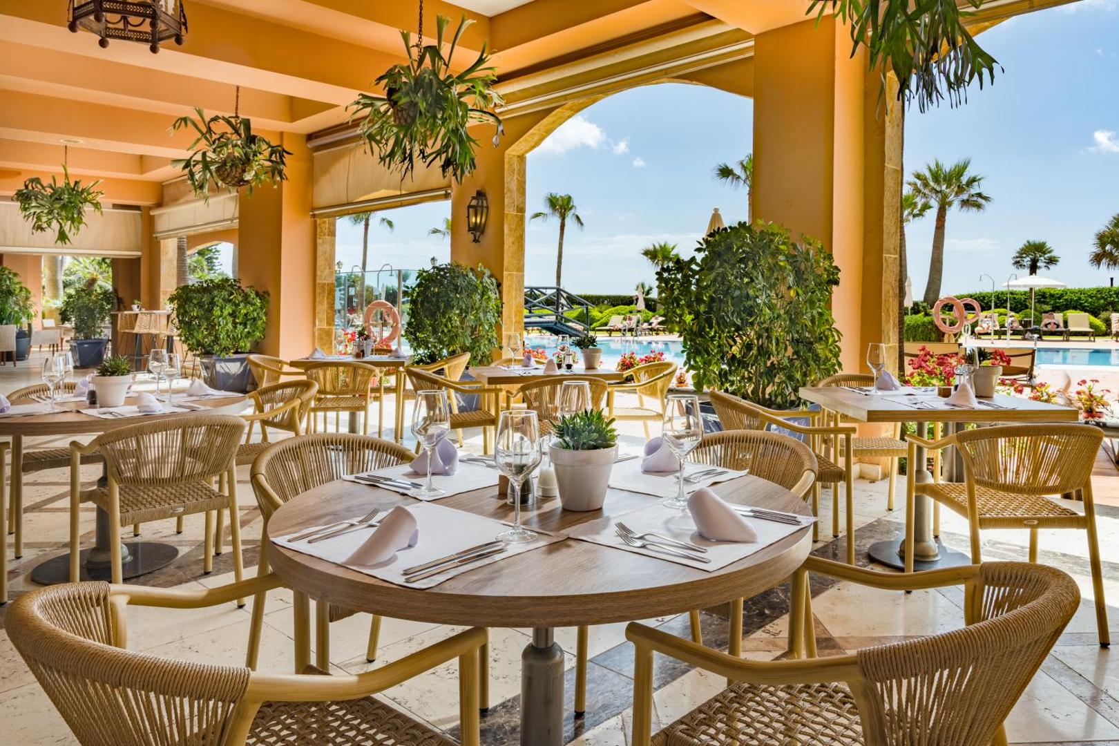 Elba Estepona Gran Hotel restorāna terase ar klātiem galdiem, pītiem krēsliem un skatu uz baseinu un dārzu