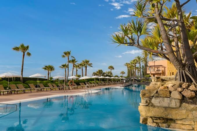 Viesnīcas Elba Estepona Gran Hotel āra baseins ar tiltiņu, eksotiskiem augiem un rindu ar saulessargiem piekrastes zonā