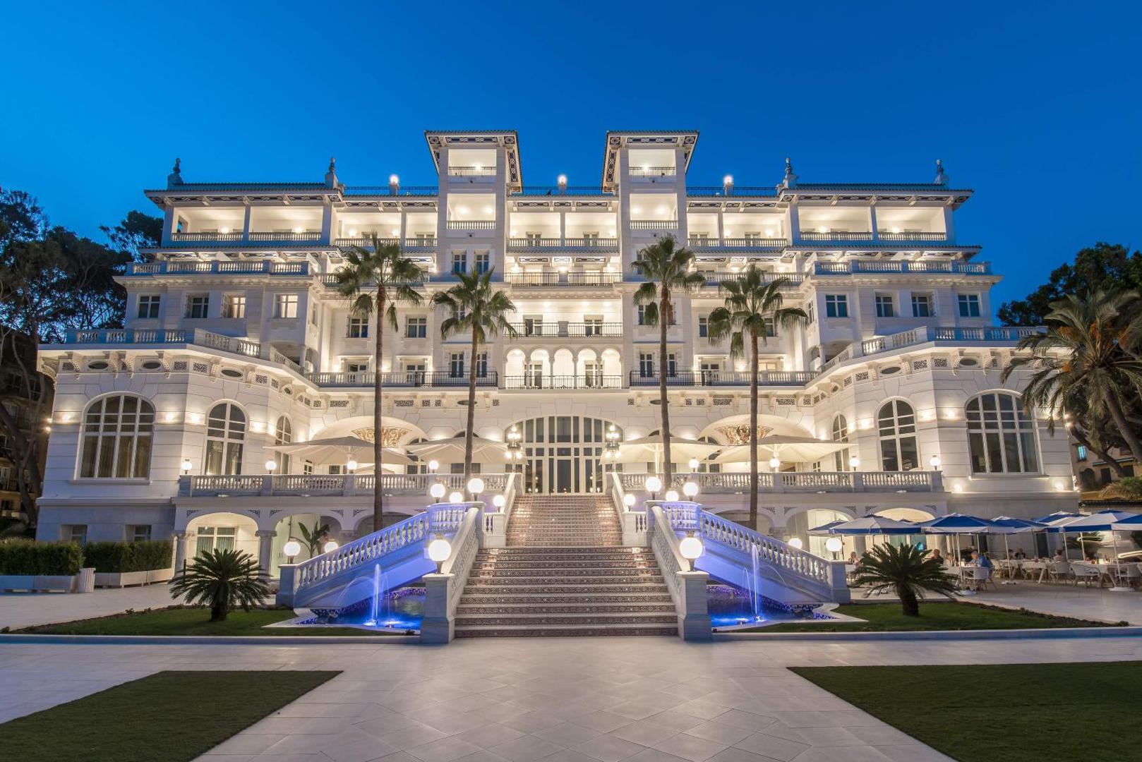 Gran Hotel Miramar fasāde vakarā Malagā, Spānijā — izgaismota vēsturiska ēka ar kāpnēm, strūklakām un palmām zilās stundas laikā