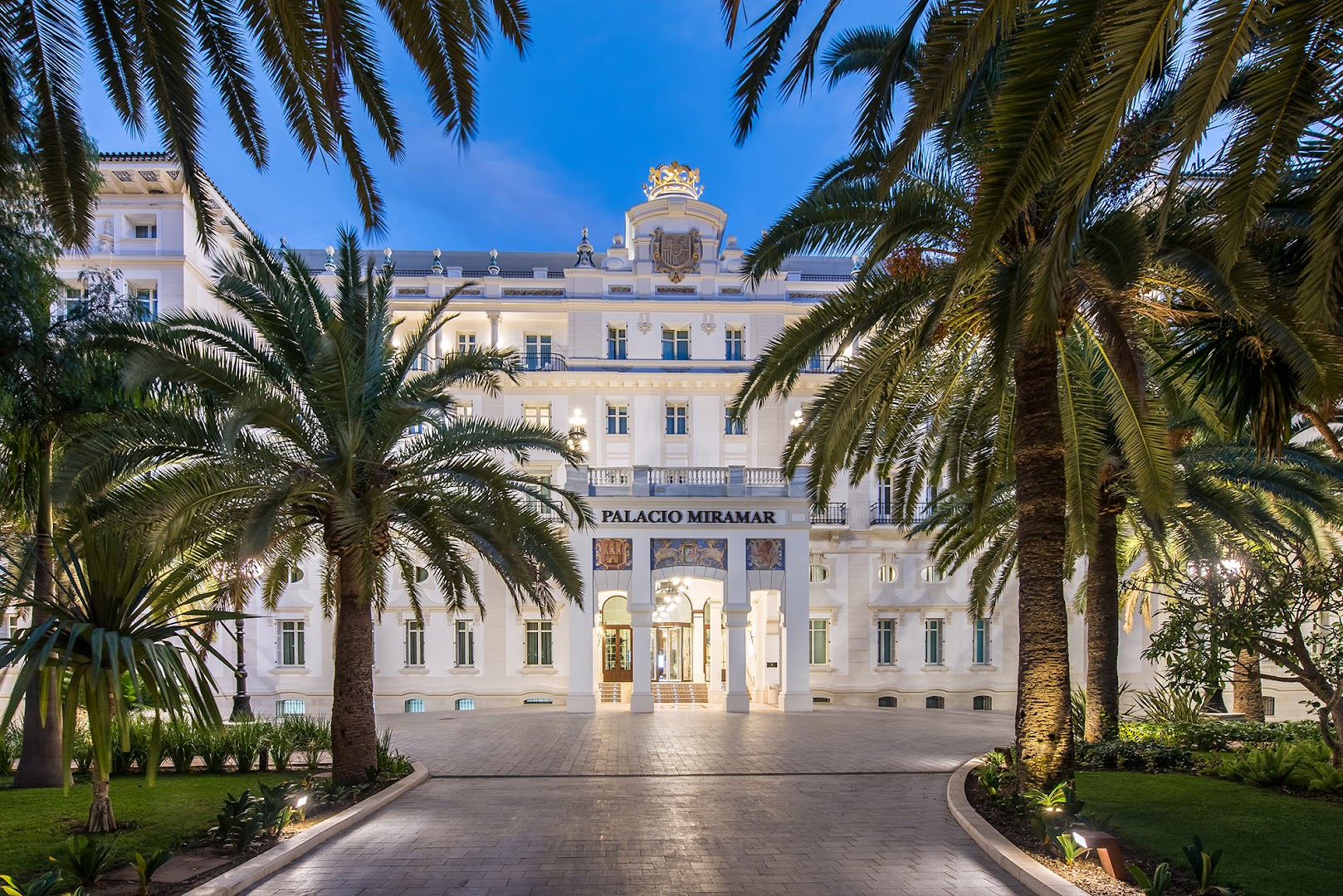 Gran Hotel Miramar galvenā ieeja caur palmu dārzu Malagā — balta pils stila ēka ar uzrakstu Palacio Miramar