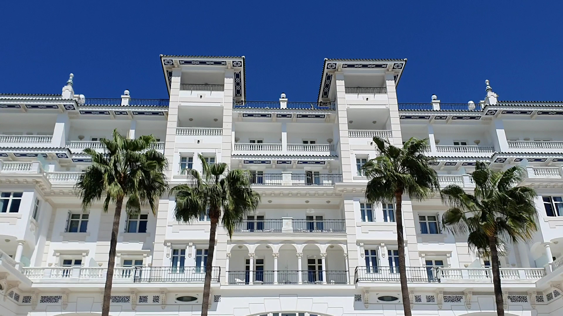 Gran Hotel Miramar augšējie stāvi un balkoni pret zilo debesu fonu Malagā — balta fasāde ar klasiskiem dekoratīviem elementiem