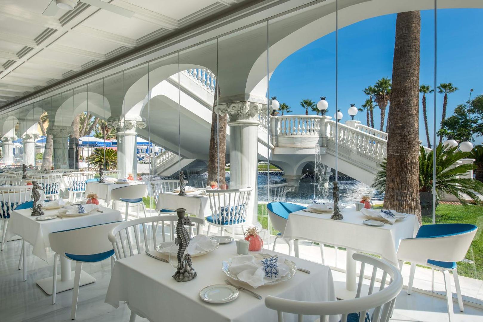 Gran Hotel Miramar elegants restorāns ar baltiem galdiem, lieliem logiem un skatu uz dārzu un kāpnēm