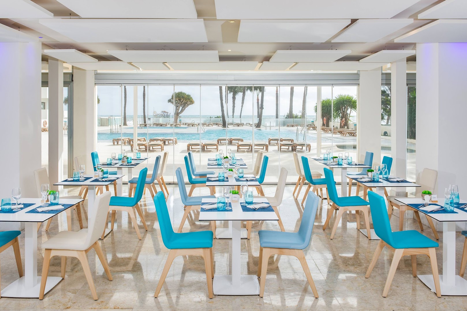 Sol House Costa Del Sol modernais restorāns ar gaiši ziliem krēsliem un skatu caur stikla sienu uz āra baseinu