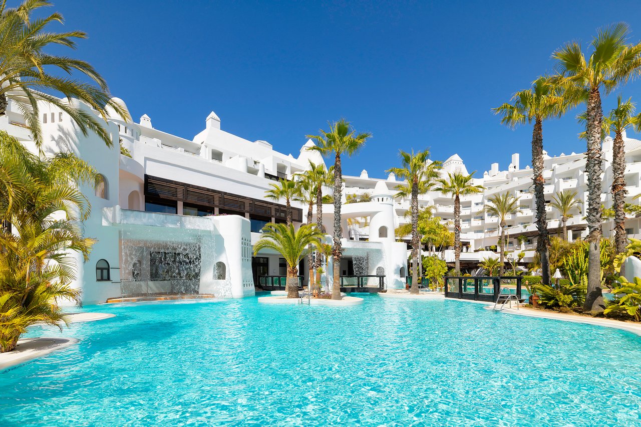 H10 Estepona Palace plašais āra baseins ar dekoratīviem ūdenskritumiem un balto viesnīcas ēku fonā saulainā dienā