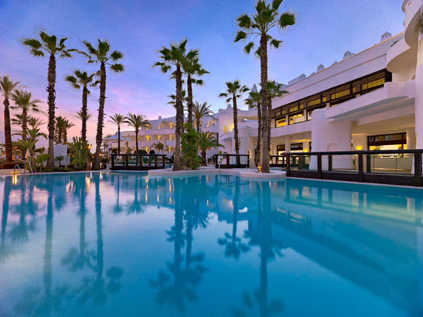 H10 Estepona Palace viesnīcas fasāde un baseins saulrieta laikā — izgaismotas palmas un balta arhitektūra piekrastē