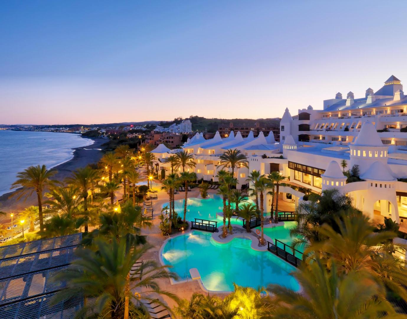H10 Estepona Palace viesnīcas skats vakarā uz izgaismotiem baseiniem, palmu dārzu un Vidusjūras piekrasti saulrieta laikā Kostadelpola reģionā.