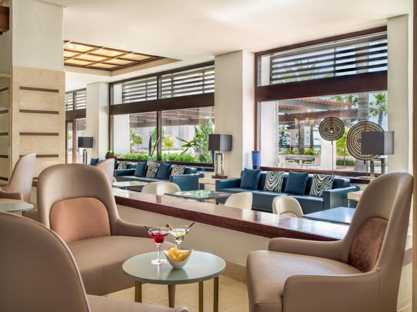 H10 Estepona Palace viesnīcas lobby bārs ar ērtiem atpūtas krēsliem, dīvāniem un skatu uz dārzu caur lieliem logiem