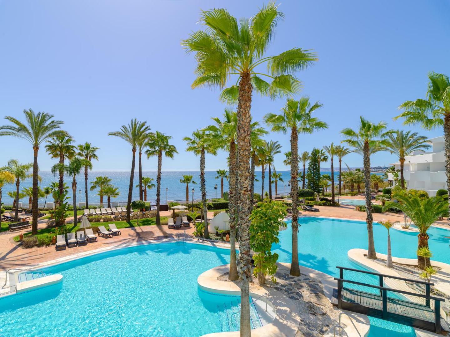 H10 Estepona Palace āra baseins ar palmām un skatu uz Vidusjūru Andalūzijā — sauļošanās zona ar atpūtas krēsliem un dārzu