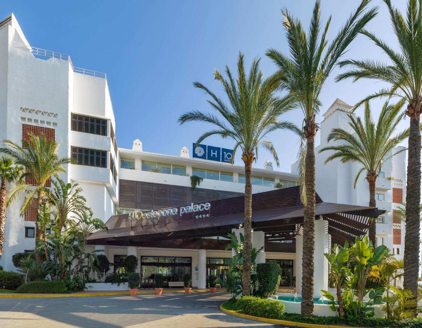 H10 Estepona Palace 4* viesnīcas galvenā ieeja ar palmām un ziedošiem augiem, demonstrējot kūrorta eleganto balto fasādi.