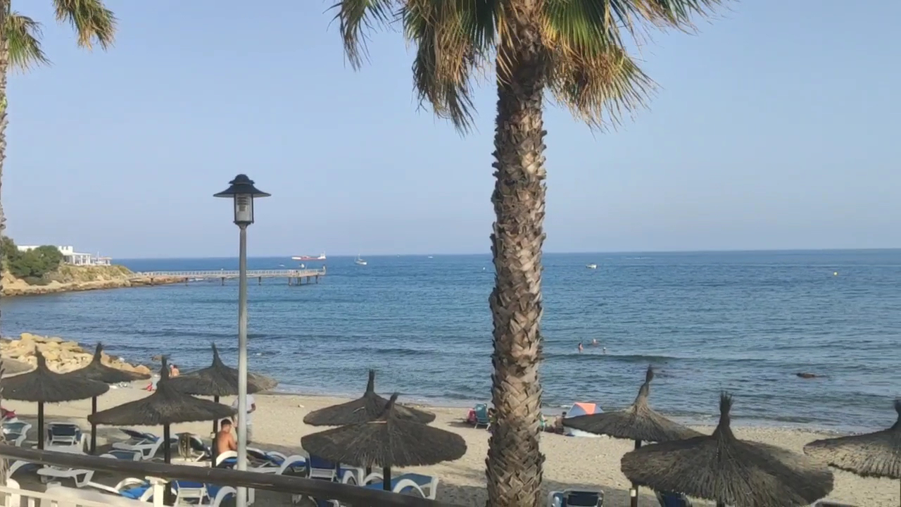 H10 Estepona Palace skats uz pludmali un Vidusjūru — smilšu krasts ar salmu saulessargiem, palmām un koka piestātni tālumā