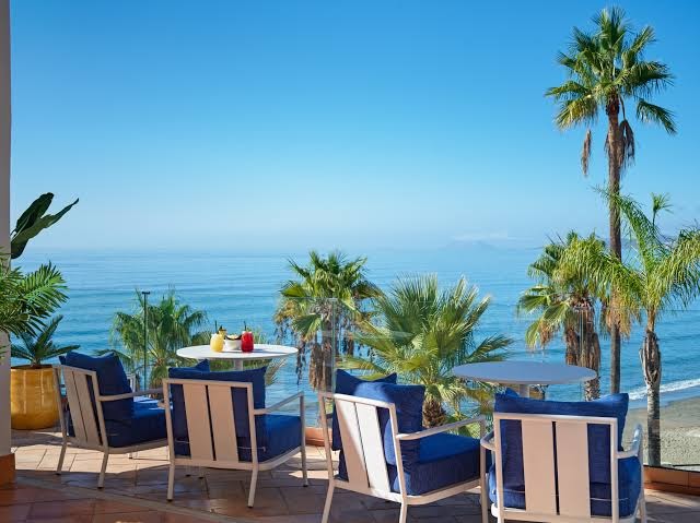 H10 Estepona Palace terase ar zilām mēbelēm un skatu uz jūru — atpūtas zona ar kokteiļiem un palmām