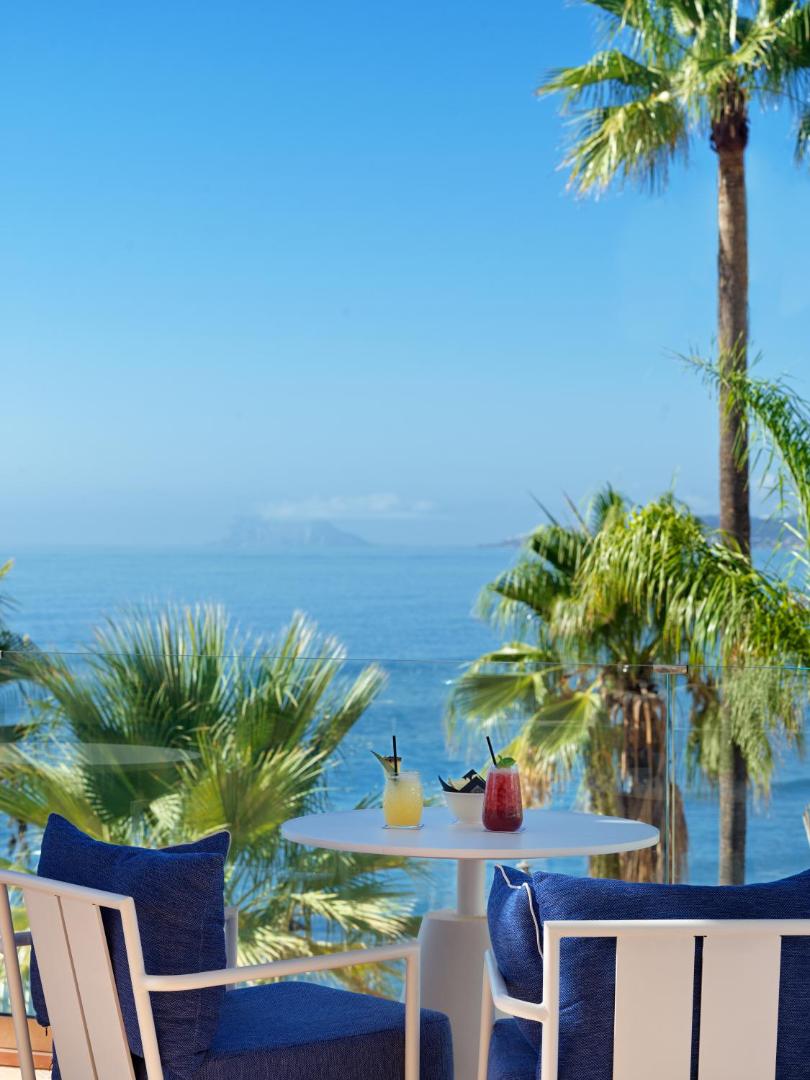 H10 Estepona Palace terase ar skatu uz Vidusjūru un Gibraltāru — balti galdiņi, zili krēsli un palmas saulainā dienā.
