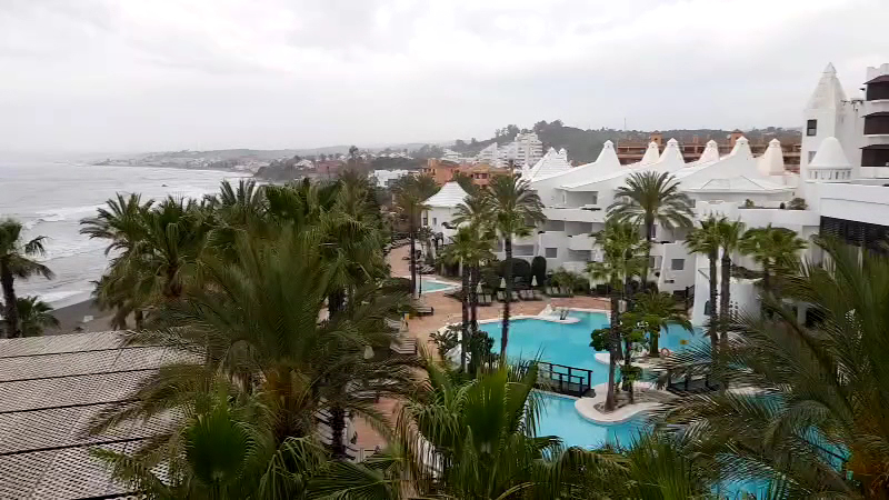 Skats no augšas uz H10 Estepona Palace baseinu zonu un piekrasti mākoņainā dienā, izceļot viesnīcas atrašanās vietu pie pašas jūras.