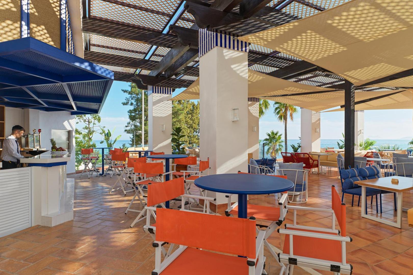 H10 Estepona Palace āra bārs un restorāns ar oranžiem krēsliem, ziliem galdiem un skatu uz jūru zem nojumes.