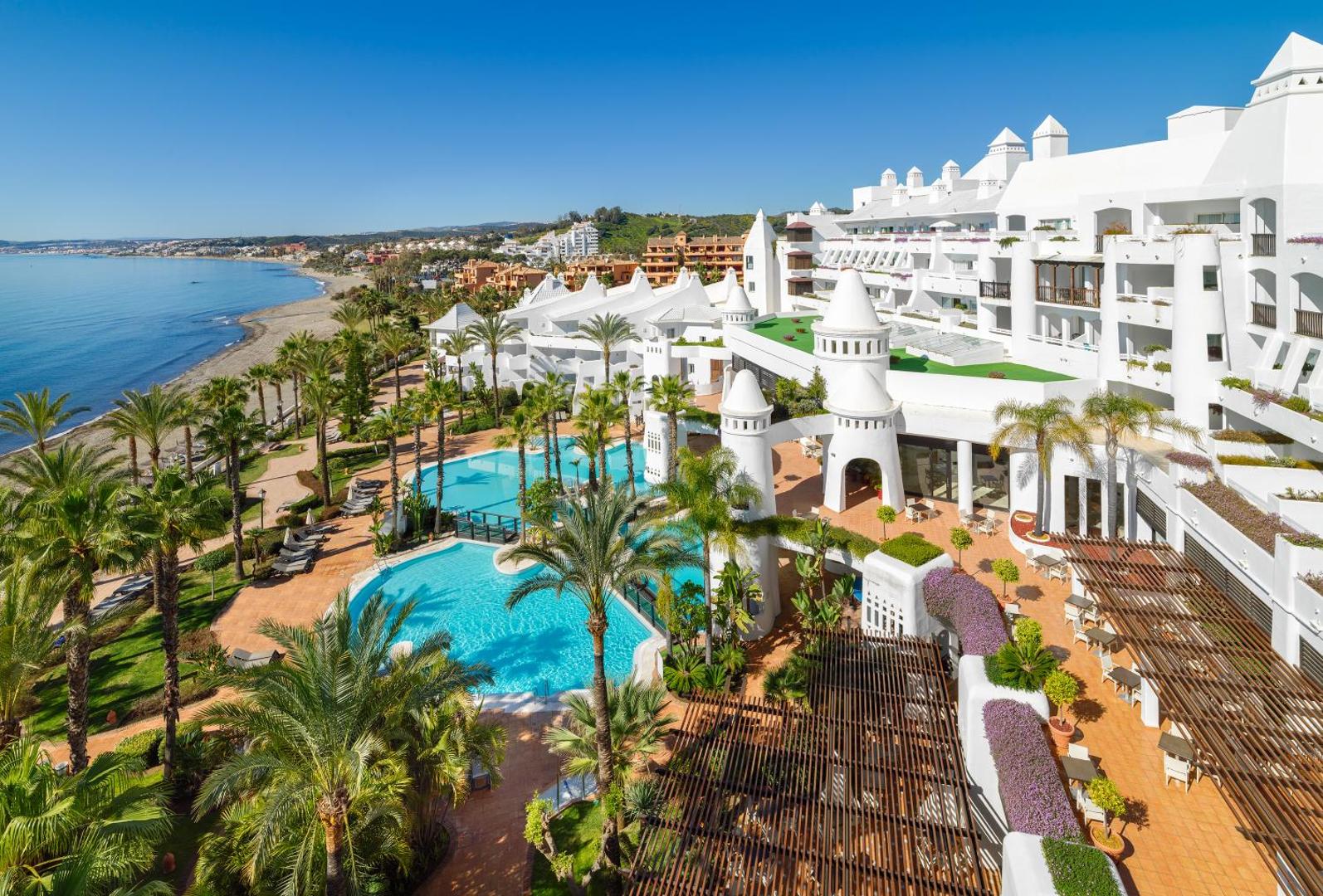 H10 Estepona Palace kūrorta arhitektūra ar baltām ēkām, vairākiem āra baseiniem un tiešu piekļuvi pludmalei saulainā dienā Esteponā.