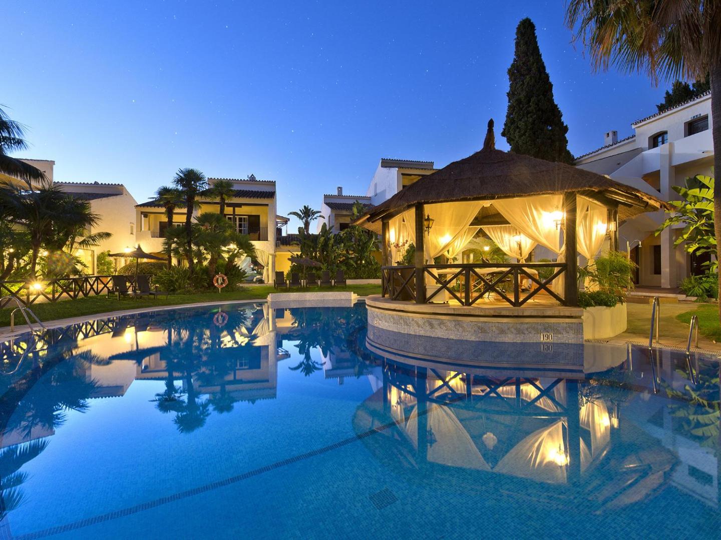 Bluebay Banus baseins un izgaismota atpūtas nojume vakarā — mājīgs apgaismojums un viesnīcas korpusi fonā