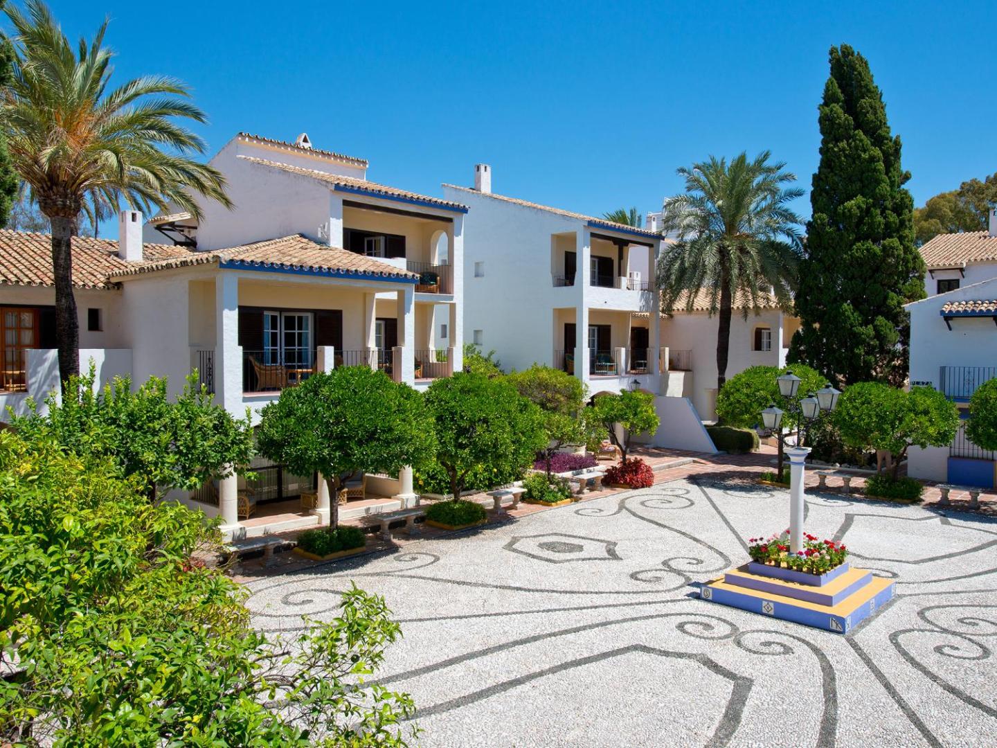 Bluebay Banus viesnīcas pagalms ar rakstainu bruģi un baltām ēkām Marbeljā, Spānijā — saulains dārzs ar palmām un citrusaugļu kokiem