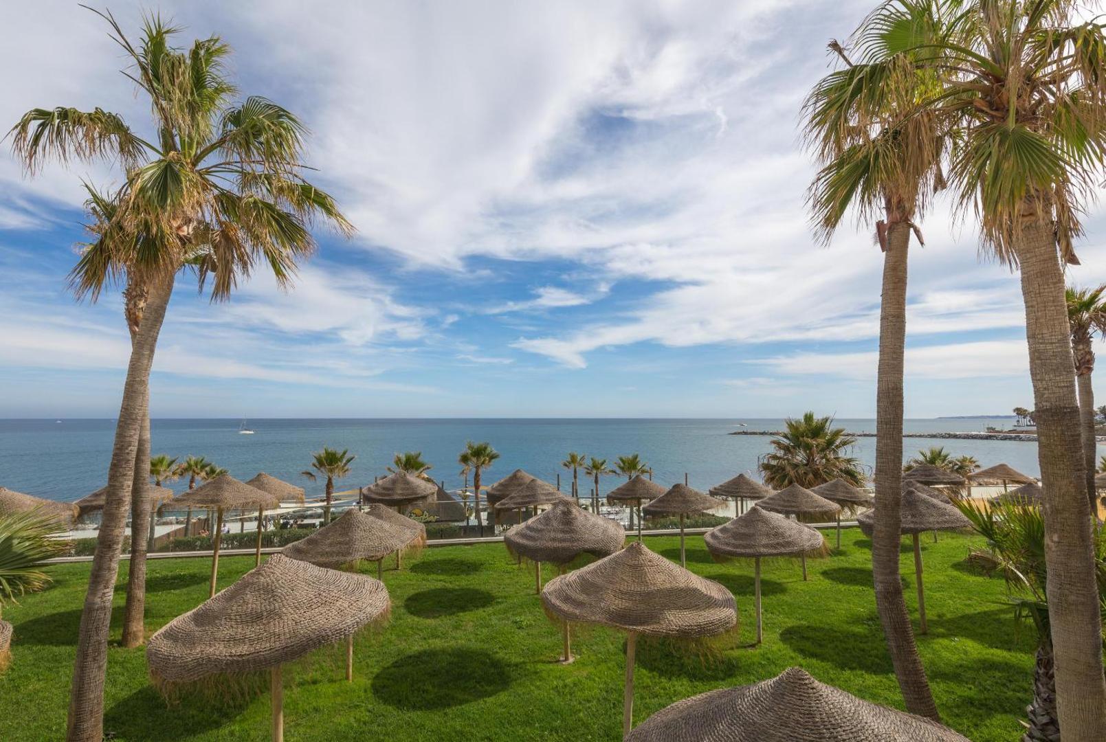 Zaļš zāliens ar salmu saulessargiem un palmām viesnīcas Best Benalmadena teritorijā, skats uz zilo jūru un debesīm.