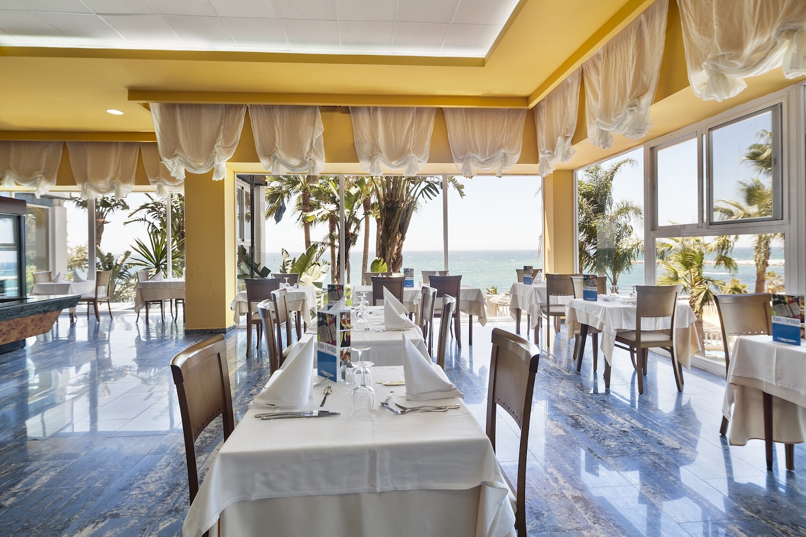Gaišs Best Benalmadena viesnīcas restorāns ar eleganti klātiem galdiem un skatu uz piekrastes palmām — plaša telpa ar dabisko apgaismojumu