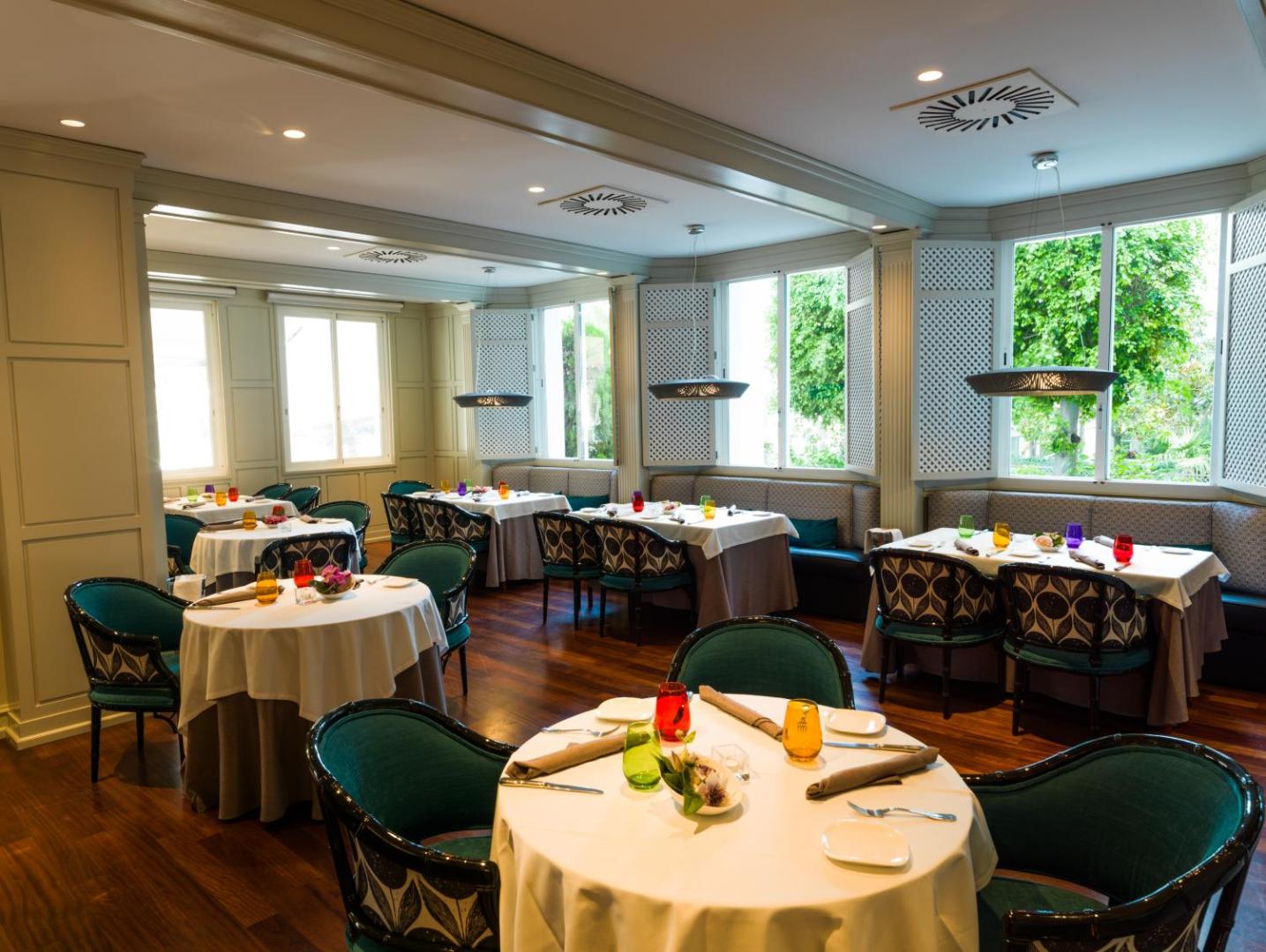 Aparthotel Monarque Sultan restorāna zāle ar eleganti klātiem galdiem, tirkīza krēsliem un lieliem logiem ar skatu uz dārzu Marbeljā.
