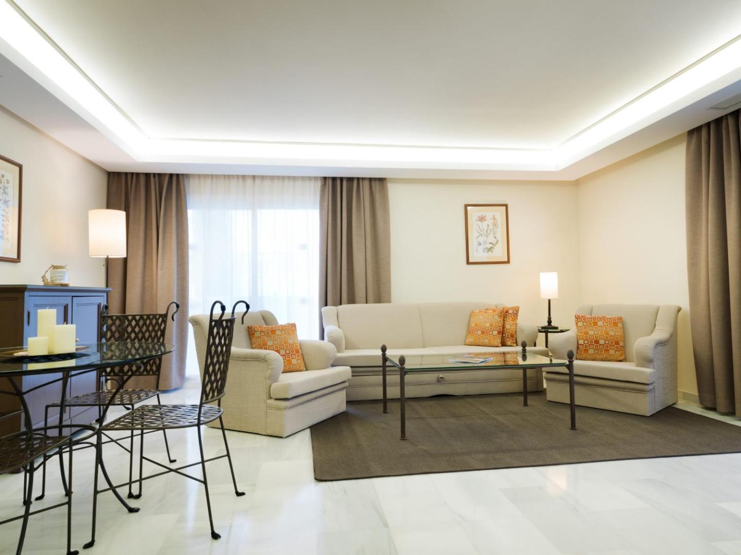 Mūsdienīgs Aparthotel Monarque Sultan numurs ar mīkstajām mēbelēm, stikla galdiņu un elegantu apgaismojumu gaišos toņos.