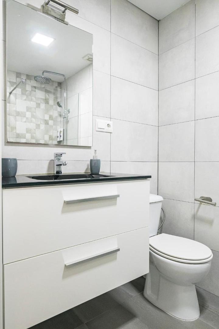 Apartamentos Mediterraneo gaiša vannasistaba ar baltu izlietnes skapi, spoguli un tualetes podu, ieturēta minimālisma stilā.