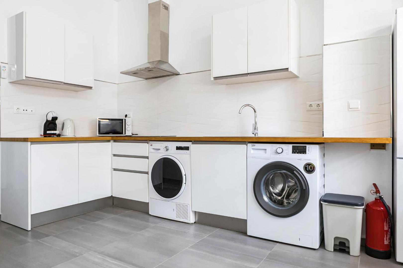 Apartamentos Mediterraneo pilnībā aprīkota virtuve ar veļas mašīnu, mikroviļņu krāsni un kafijas aparātu — balti skapīši un koka virsma