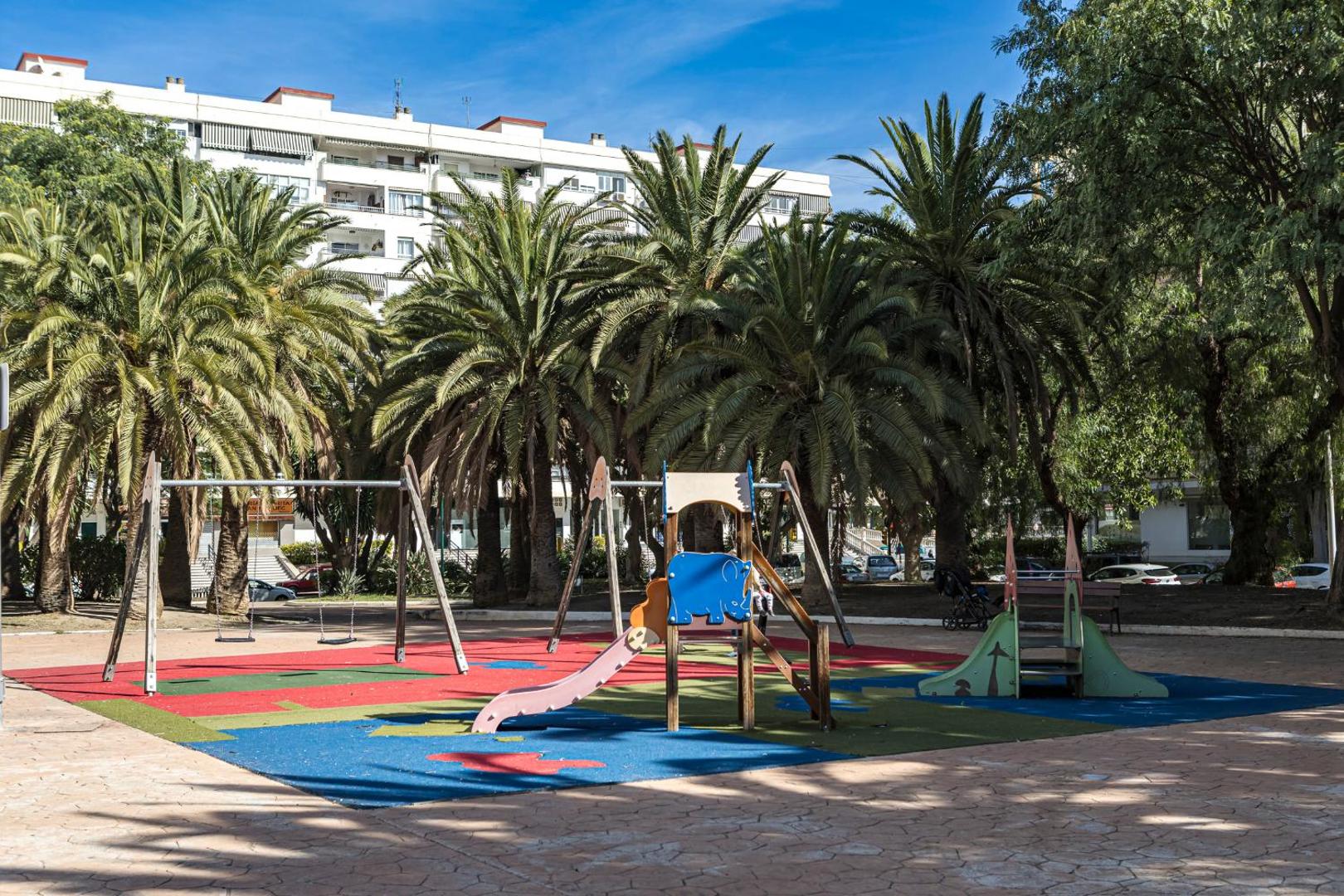 Apartamentos Mediterraneo bērnu rotaļu laukums ar slidkalniņiem un šūpolēm, ko ieskauj augstas palmas un pilsētas apstādījumi.
