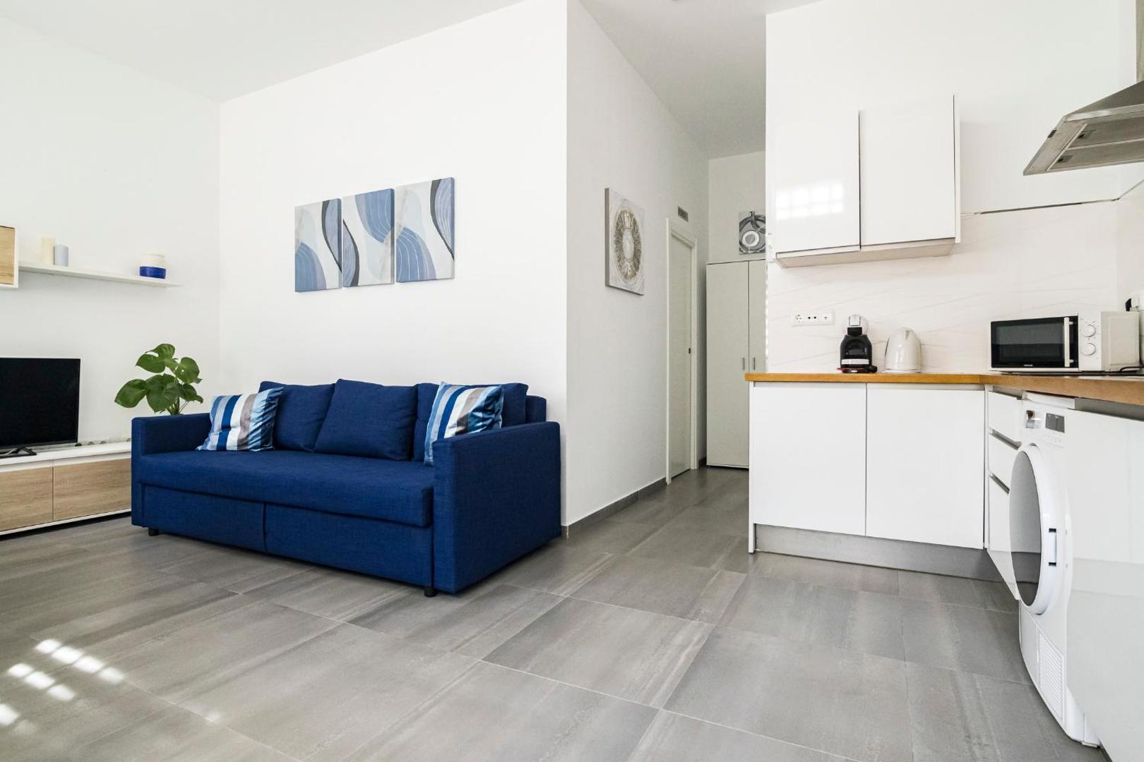 Apartamentos Mediterraneo studijas tipa numura skats uz dzīvojamo zonu un virtuves stūri — balts interjers ar ziliem akcentiem