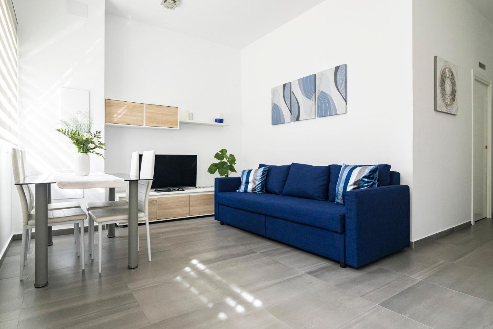 Apartamentos Mediterraneo gaiša dzīvojamā zona ar zilu dīvānu, ēdamgaldu un TV — mūsdienīgs interjers ar pelēkām flīžu grīdām