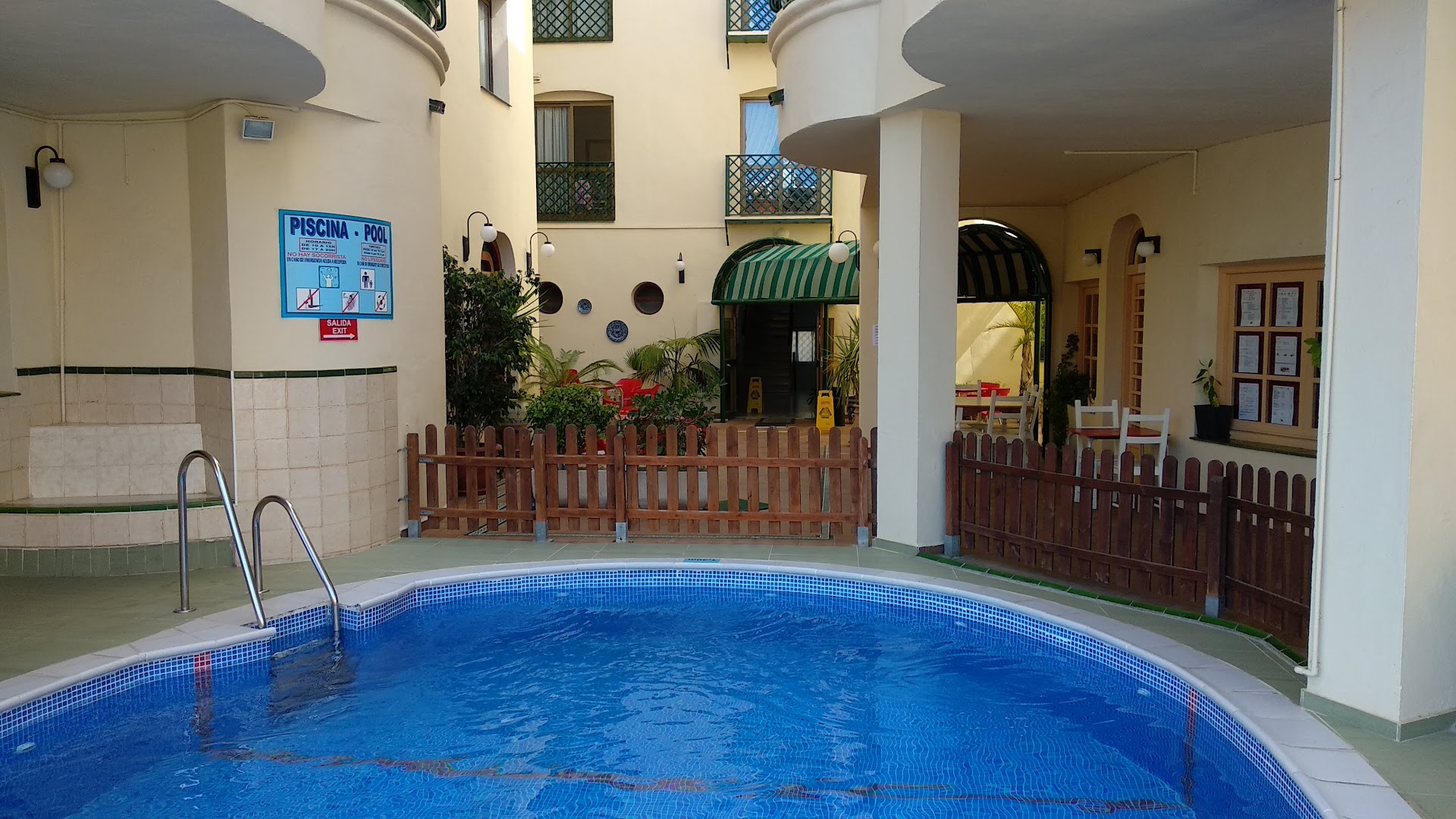 Apartamentos Mediterraneo iekšpagalma baseins ar zilu mozaīkas apdari, koka sētu un ieeju viesnīcas koplietošanas telpās Spānijas piekrastē.