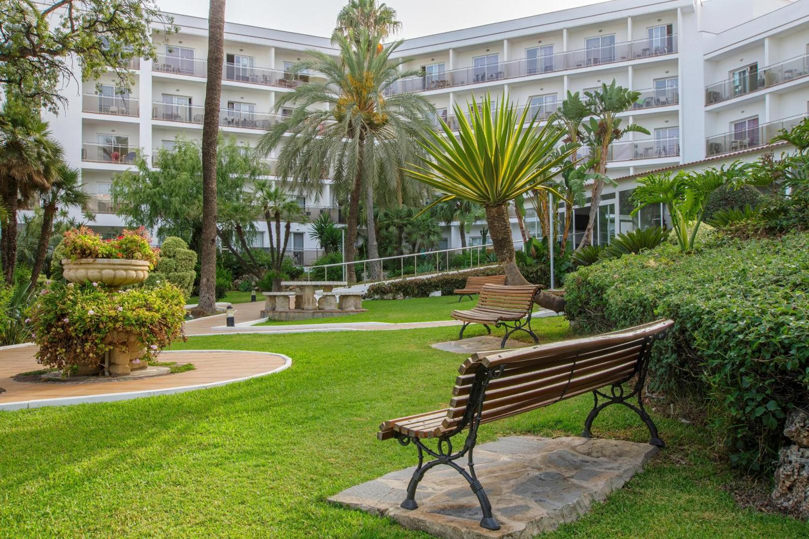 Atpūtas zona Hotel Best Siroco dārzā ar koka soliņiem, zālāju un palmām pie viesnīcas korpusa Benalmadenā