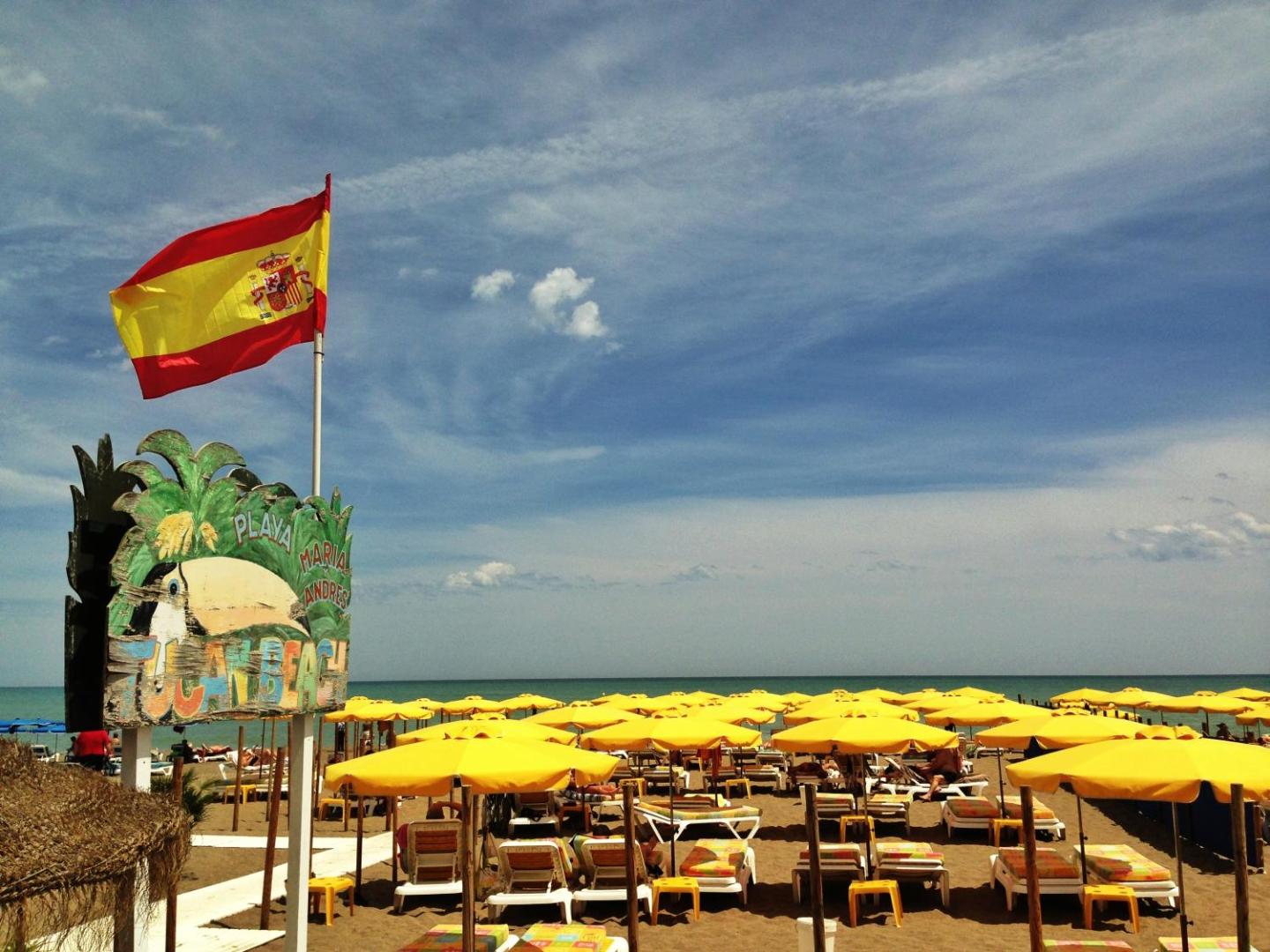 Playa Maria Andres pludmale pie Carmen Teresa viesnīcas — dzelteni saulessargi, Spānijas karogs un Tucan Beach zīme smiltīs.