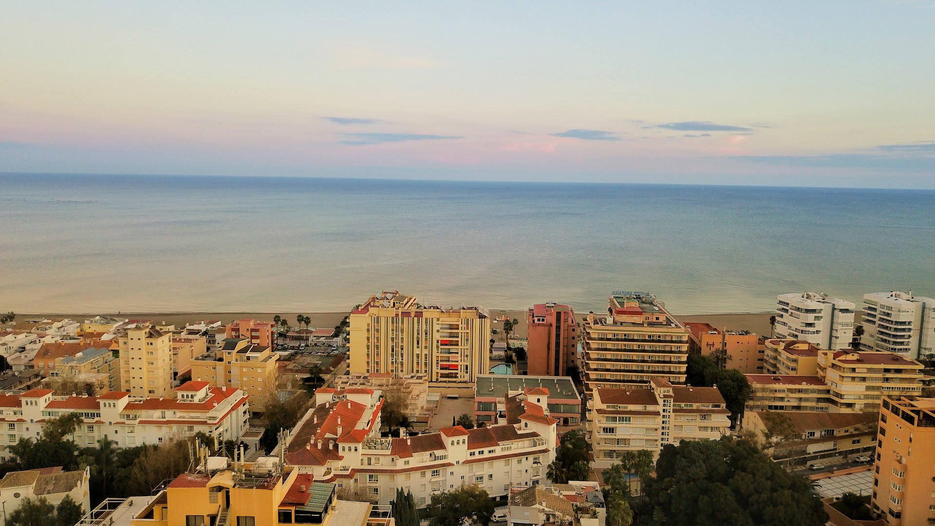 Carmen Teresa viesnīcas apkārtnes panorāma Torremolinosā saulrietā — pilsētas apbūve pie Vidusjūras krasta līnijas zem sārtām debesīm.