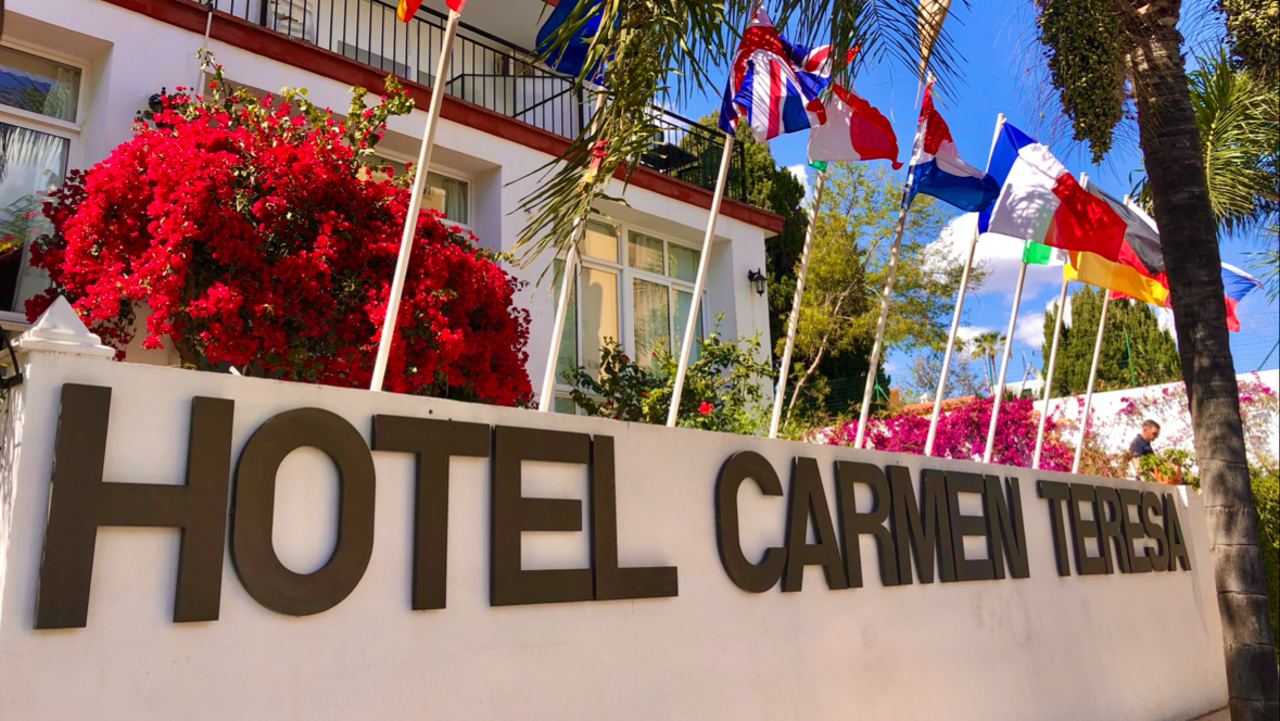 Hotel Carmen Teresa ieeja ar uzrakstu uz baltas sienas, krāšņiem sarkaniem ziediem un dažādu valstu karogiem