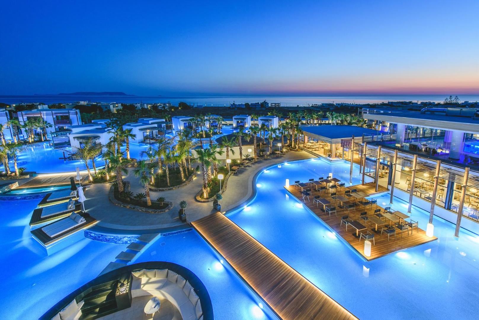 Stella Island Luxury Resort & Spa vakara apgaismojums pie galvenā baseina un restorāna terases ar skatu uz jūru saulrietā