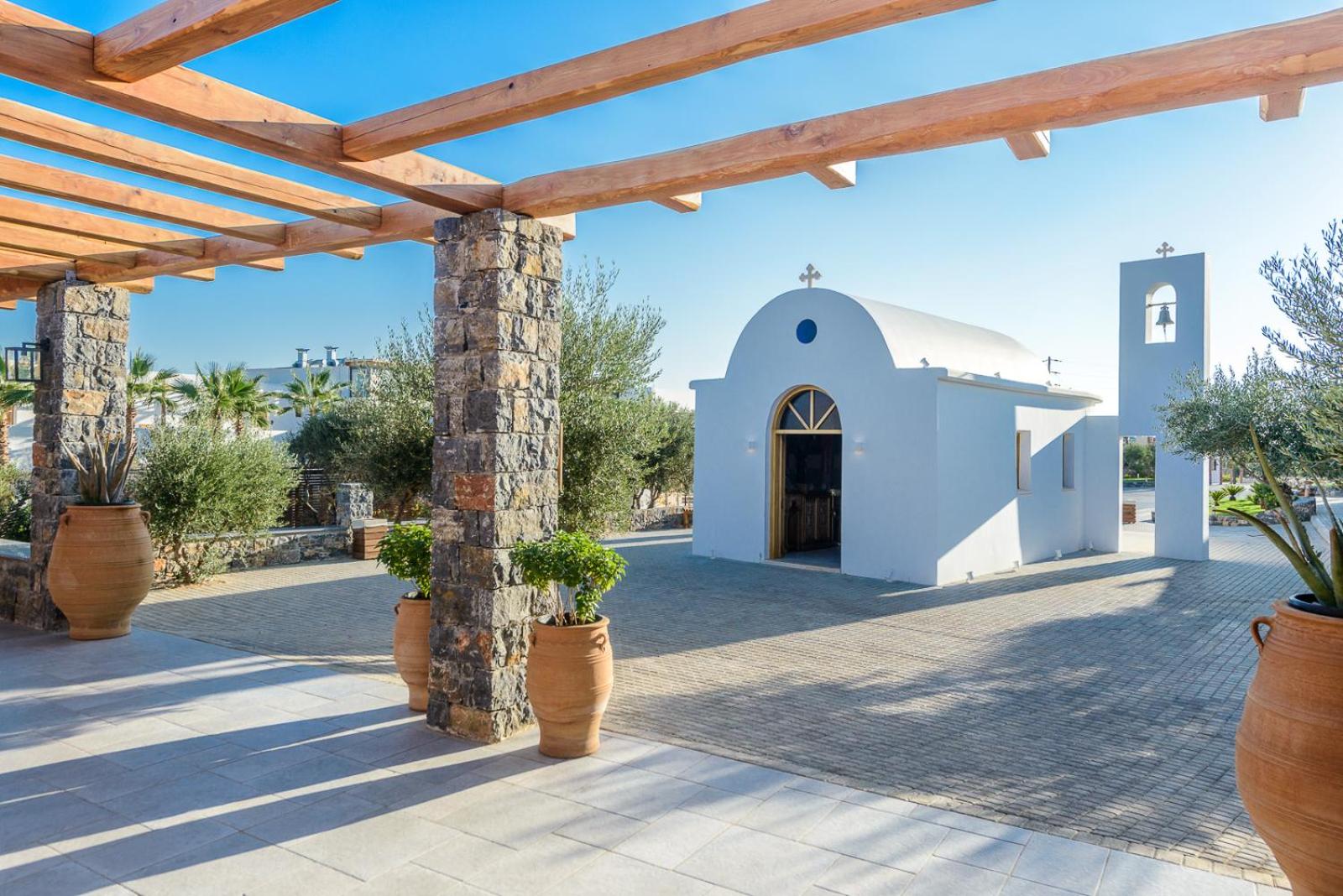 Stella Island Luxury Resort & Spa balta grieķu stila kapela ar zvanu torni kūrorta teritorijā zem koka pergolas saulainā dienā