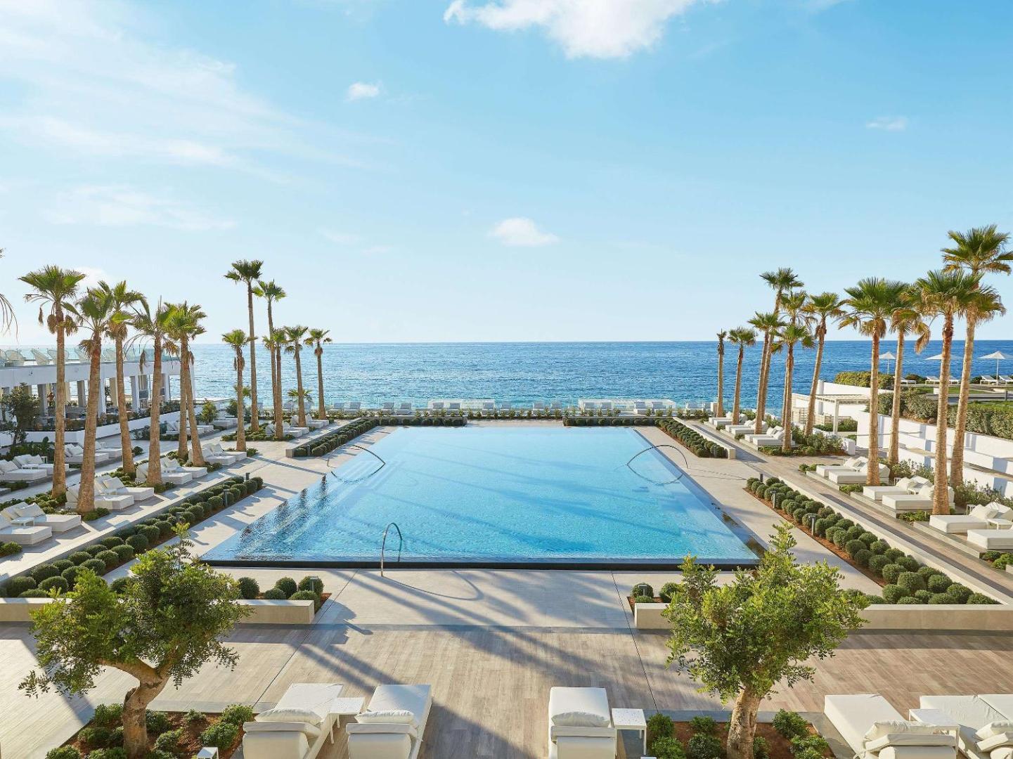Grecotel Lux Me White Palace galvenais āra baseins ar palmām un skatu uz jūru — plaša sauļošanās zona ar gultām