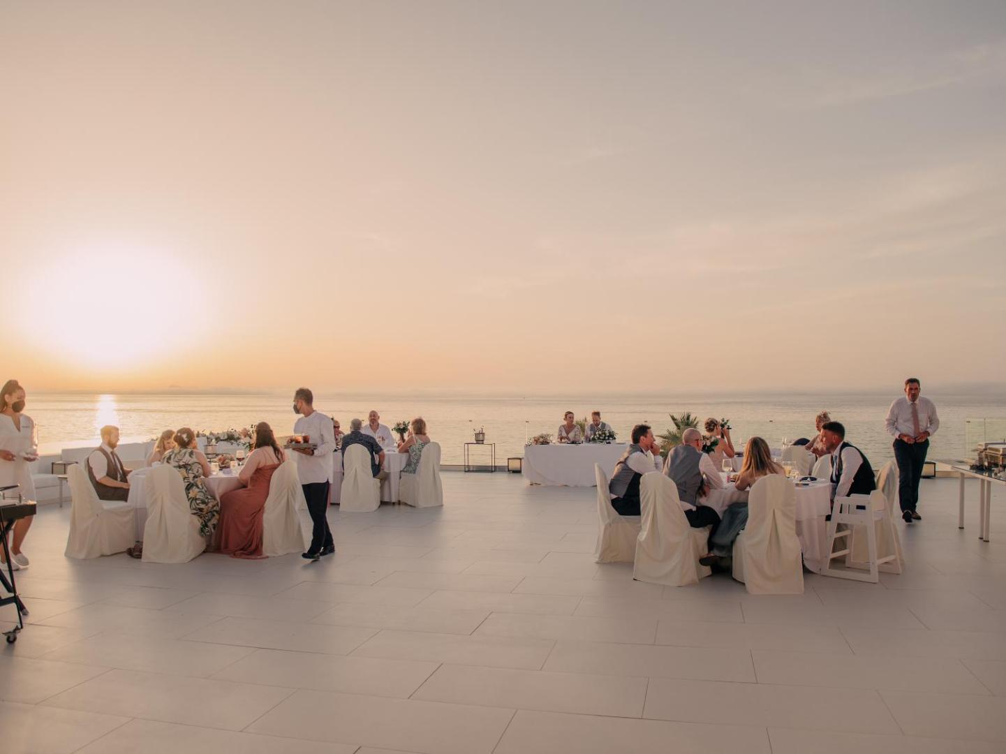 Grecotel Lux Me White Palace āra terase ar svētku galdiem un viesiem saulrieta laikā pie jūras Krētā