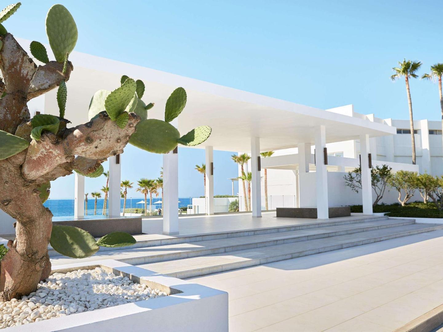 Grecotel Lux Me White Palace ieeja ar modernu arhitektūru, baltām kolonnām, kaktusu un palmām Krētas piekrastē