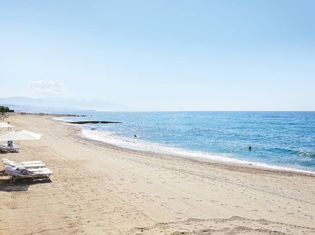 Grecotel Lux Me White Palace plašā smilšu pludmale Krētā ar saulessargiem, sauļošanās krēsliem un skatu uz dzidri zilo jūru